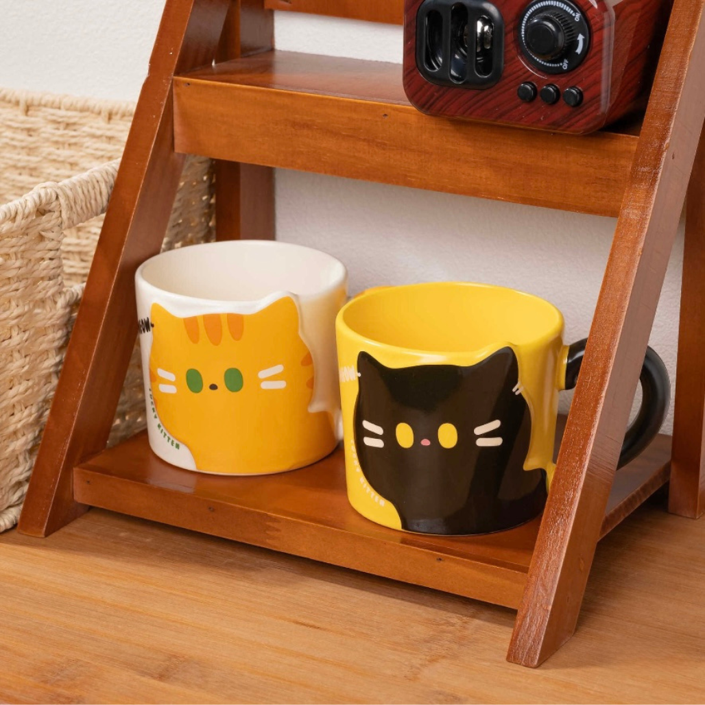 Two cat-themed mugs on a wooden shelf with a radio in the background.