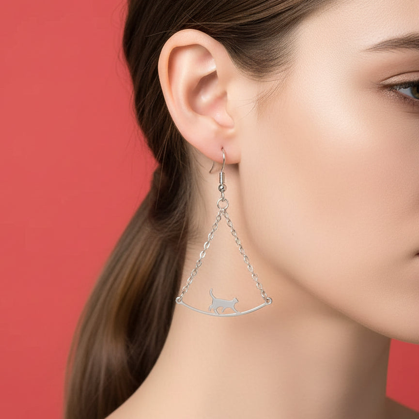 Silver earrings feature a cat precariously walking across a bridge suspended by two delicate chains on a dark background