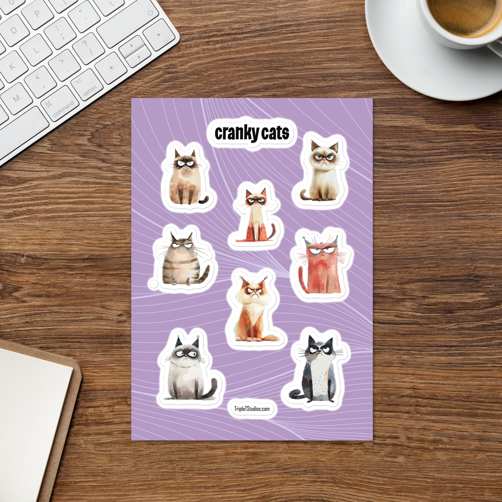 A sheet of die-cut stickers featuring illustrated grumpy cats on a lavender background, placed on a wooden desk with a keyboard, coffee cup, and notebook partially visible around it.