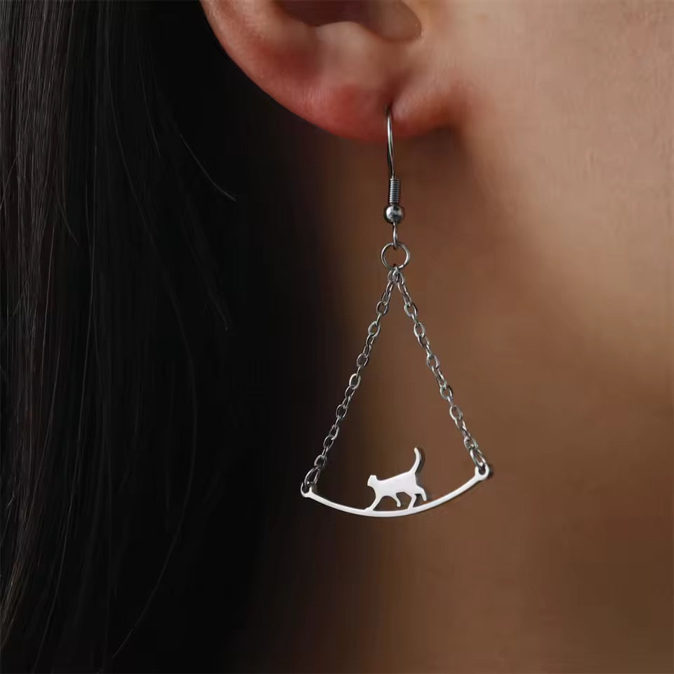 Silver earring with a cat design hanging from a chain, worn on an ear.