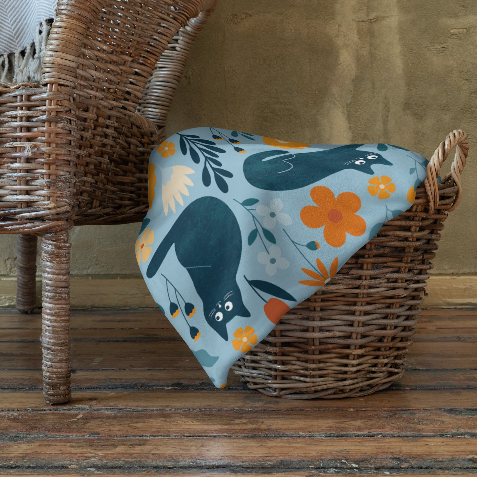 Cozy fleece blanket with blue cats and flower pattern draped over a wicker basket on a wooden floor.