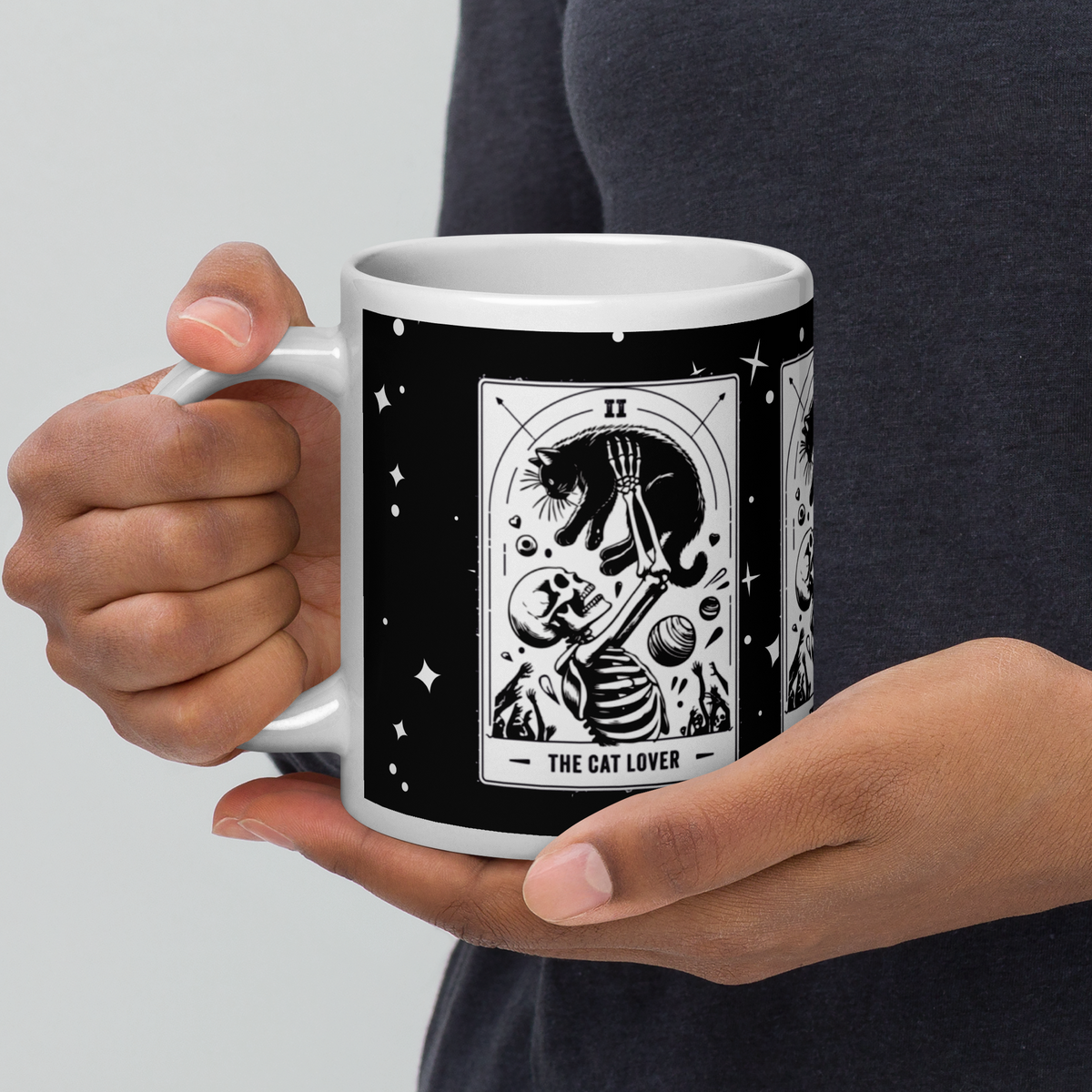 A person holding a white ceramic mug with a black wraparound graphic featuring a tarot card illustration labeled 'THE CAT LOVER.' The person is wearing a dark gray shirt against a light gray studio background.