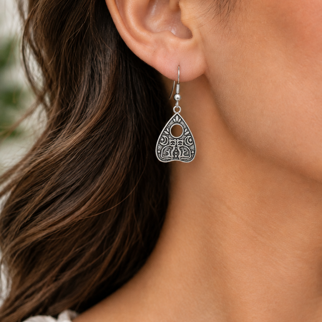 Silver metal earrings that look like a ouija board planchette hang from fish hook earring wires. Two cats and moons are in the design