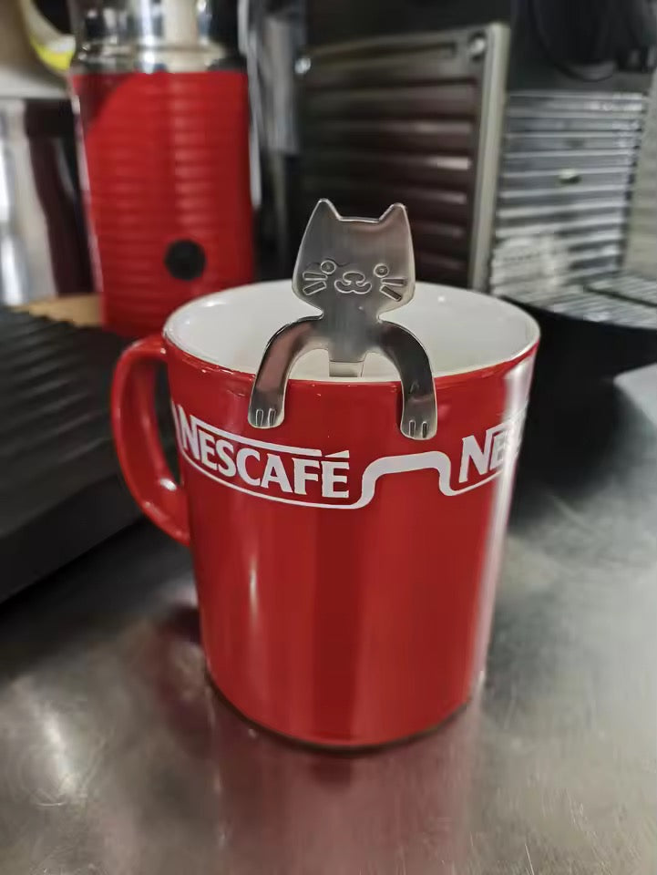 Red Nescafe mug with a cat-shaped spoon on a metallic surface near coffee machines