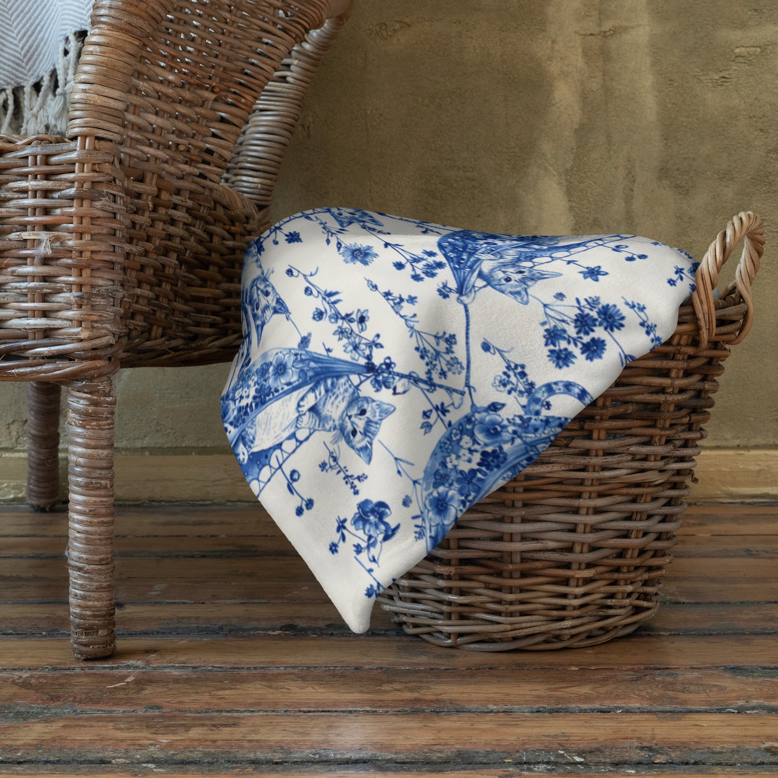 Blue and white Triple T Studios Cats in Hammocks floral patterned blanket draped over a basket on a wooden floor.