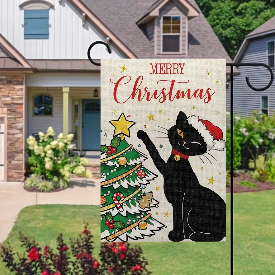 A decorative garden flag hanging on a black metal stand in a front yard. The flag has a cream background with the words “MERRY Christmas” in red script, a black cat wearing a red Santa hat, and a decorated Christmas tree with gold stars.