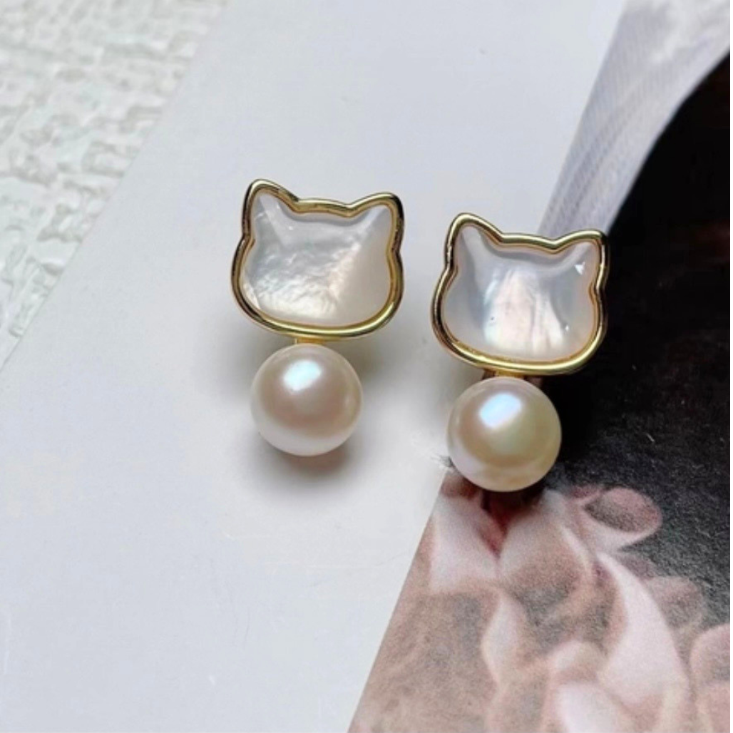 Cat Head shaped earrings with Mother of Pearl inset and a freshwater pearl drop sit on a desktop surface.
