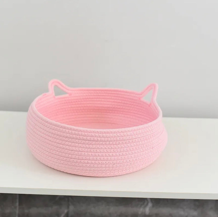 A light pink Cotton Rope basket with cat-ear handles, sitting on a white shelf against a light gray wall.