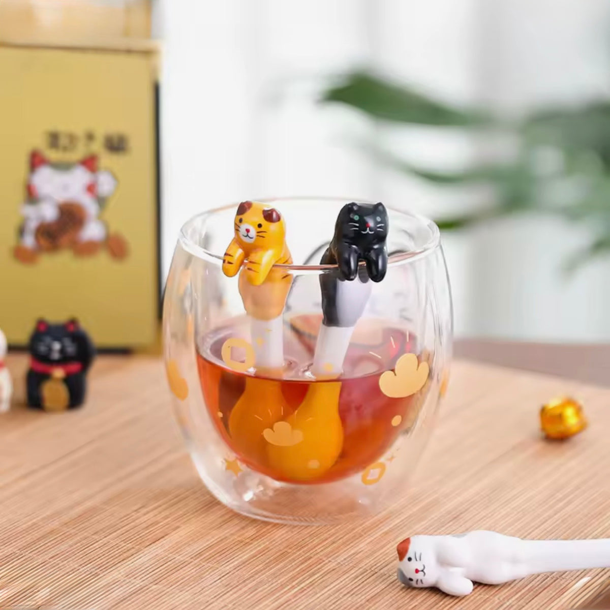 Glass cup with ceramic cat spoons that dangle over edge and other cat-themed decorations on a wooden surface