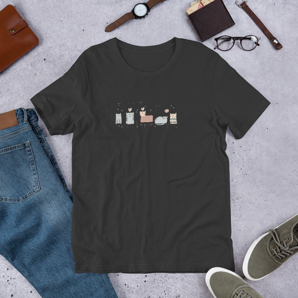Dark grey t-shirt with abstract SQUARE CATS graphic, surrounded by jeans, shoes, and accessories on a concrete surface.