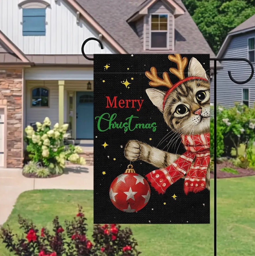A decorative garden flag hanging on a black metal stand in a front yard. The flag features a tabby cat wearing a red reindeer antler headband and a red knit Christmas scarf, holding a red ornament against a black background with the words 'Merry Christmas' in red and green.