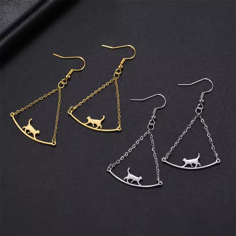 Gold and silver earrings feature a cat walking on a bridge suspended by two chains. Earrings have french wires. Background is a dark surface