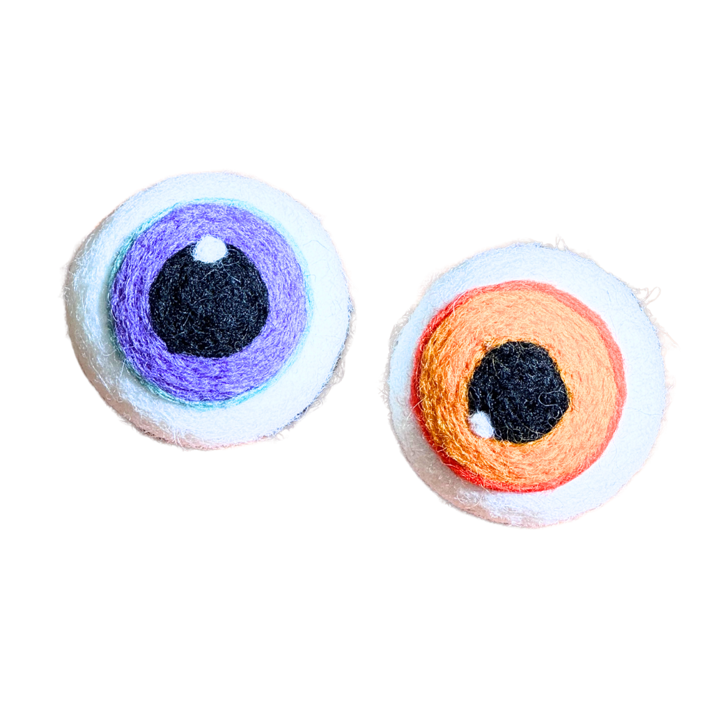 Two handmade felt wool eyes on a plain white background, one with purple and blue details and one with orange and red details.