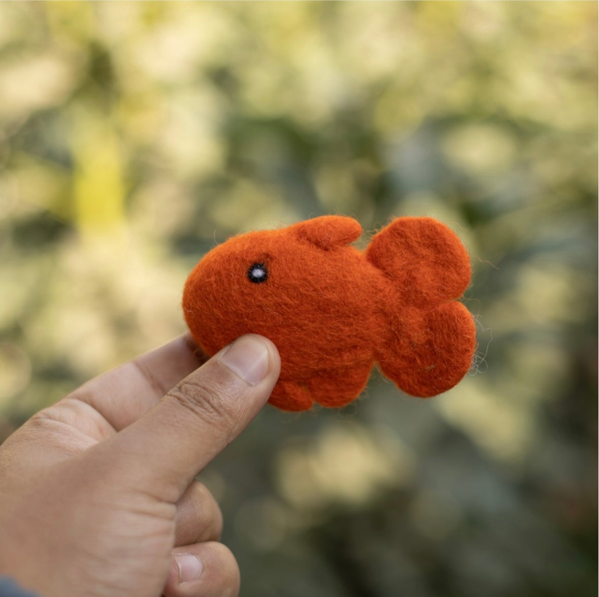 Hand holding a small orange felt fish cat toy against a blurred natural background
