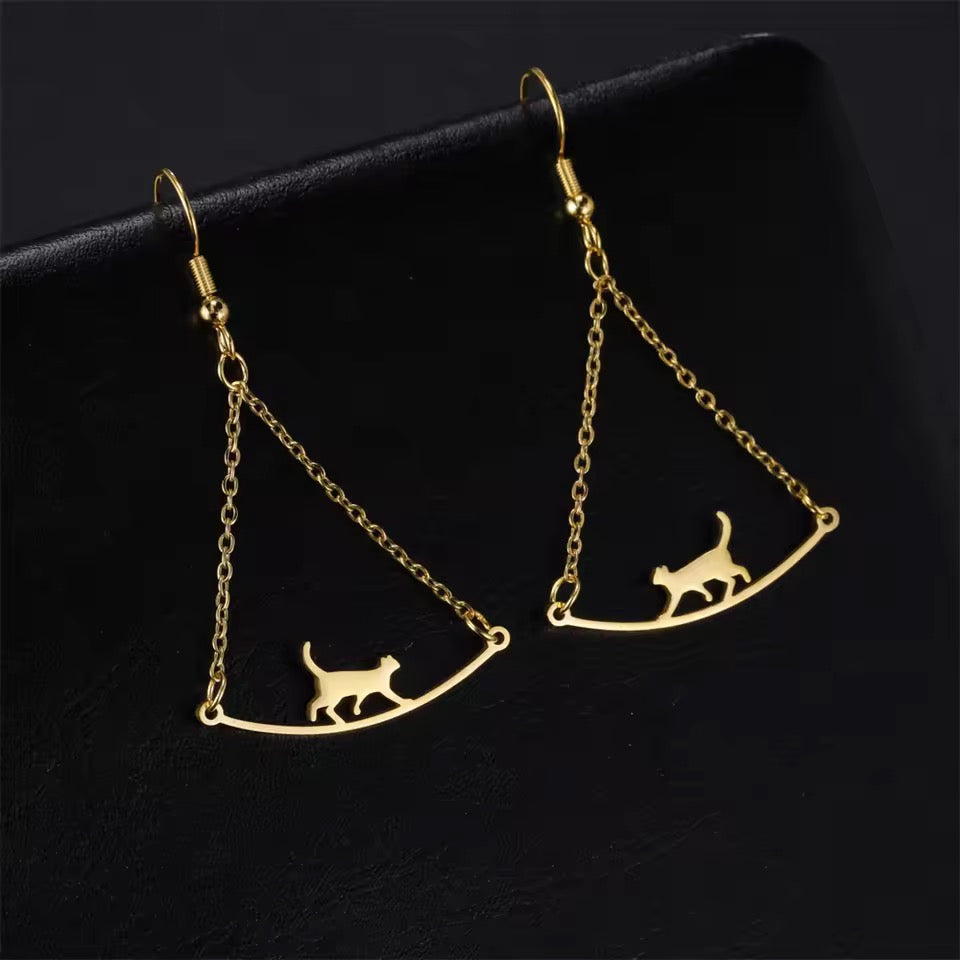 Triple T Studios Cat Walk goldentone earrings featuring a cat walking across a bridge suspended precariously by two chains on a black background