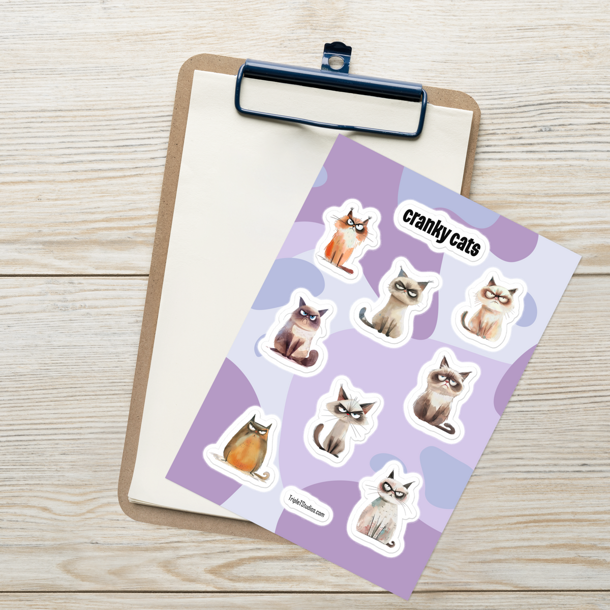 A sheet of die-cut stickers featuring illustrated cats with angry expressions on a lavender patterned background, placed on top of a clipboard over a light wood surface. The sheet shows the text 'crankycats' and TripleTStudios logo.