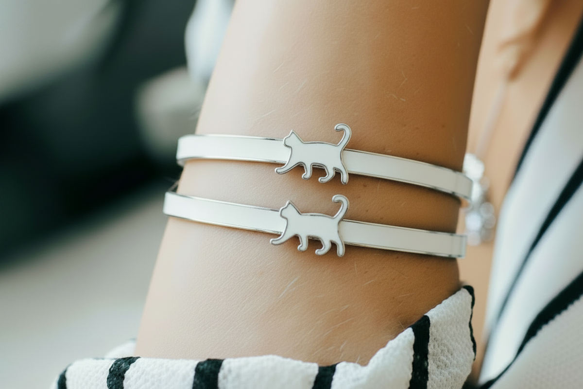 A close-up of a person's wrist wearing two slim white enamel bangle bracelets with walking white cats.
