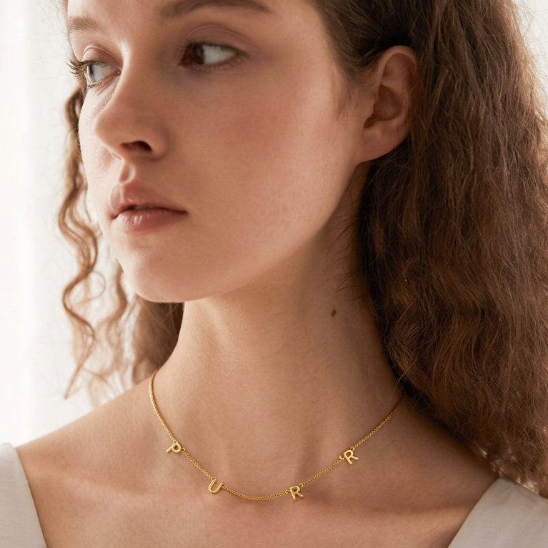 A close-up of a woman wearing a delicate gold chain necklace with small capital letters spelling 'PUR' in gold.